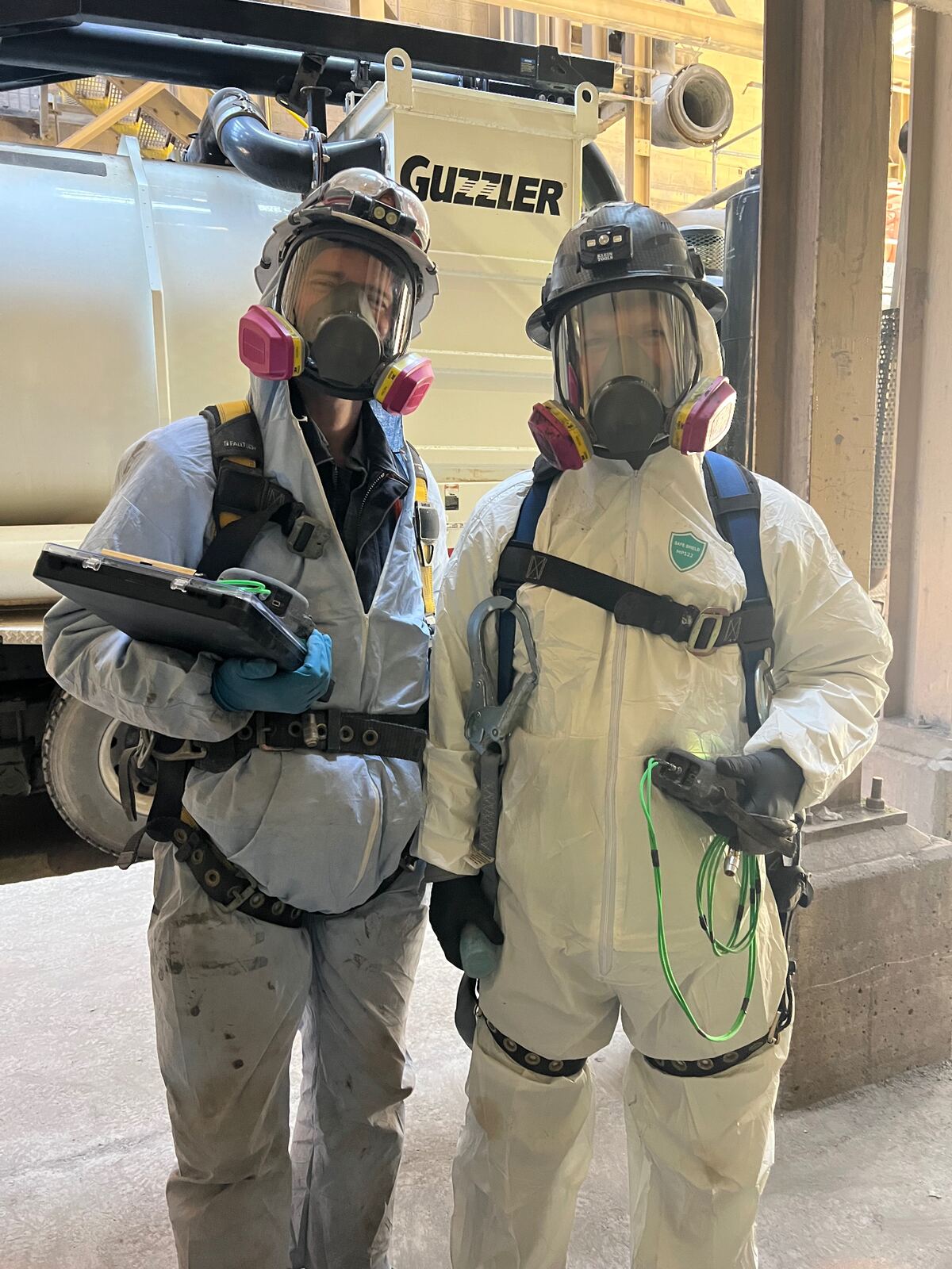 Two AIT inspectors in full PPE with respirators, harnesses, and inspection tablet at an industrial worksite