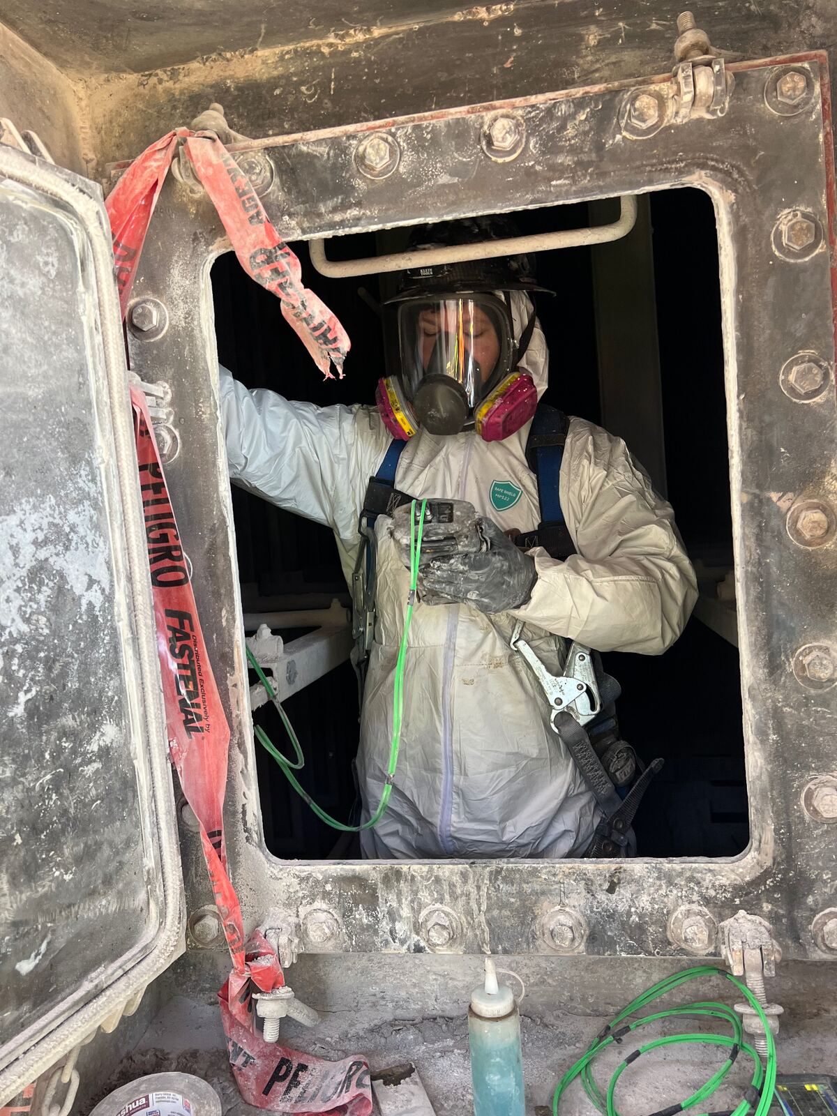 AIT technician in Tyvek suit, full-face respirator, and fall-arrest harness exiting a bolted vessel manway