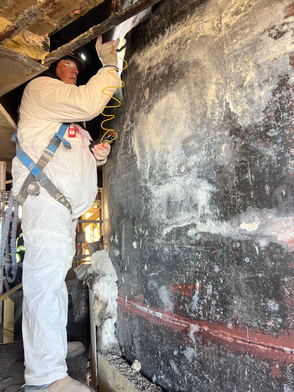 AIT inspector performing ultrasonic thickness scan on a corroded vessel shell from scaffolding